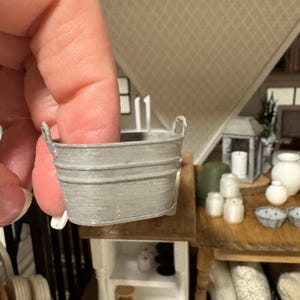 May include: A miniature gray metal tub with a ribbed exterior and two small handles on the sides. The tub has a smooth, matte finish and a slightly tapered oval shape, designed for use in a dollhouse or as a small decorative accessory.