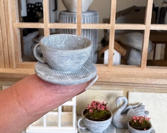 May include: A miniature gray teacup and saucer set, held by a finger, with a textured surface. The teacup has a handle and sits atop a matching saucer. Other miniature decorative items are in the background.