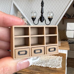 Può includere: Un'unità a scaffale in legno in miniatura con nove scomparti e tre cassetti. I cassetti hanno maniglie in metallo nero. L'unità a scaffale è realizzata in cartone marrone e ha un aspetto rustico.