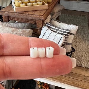 Peut inclure: Trois petites bougies blanches en forme de pilier avec des mèches noires. Les bougies sont tenues dans une main.