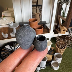 May include: Two miniature grey ceramic vases. One is a rounded, textured vase with a narrow neck. The other is a small, round pot. The vases are held in a person's hand, with other miniature decor in the background.