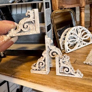 Può includere: Primo piano di staffe angolari bianche e ornate in miniatura con dettagli a volute. Le staffe sono invecchiate con una finitura anticata. Altri mobili in miniatura sono sullo sfondo, suggerendo una casa delle bambole o un progetto artigianale.