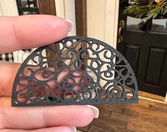 May include: Black metal half-circle decorative doormat with a delicate, intricate design. The doormat is made of a thin, lightweight material and has a slightly curved shape.