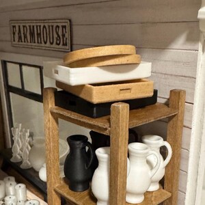 May include: A wooden shelf unit with three shelves, holding a stack of three trays, two white and one brown, and a collection of white and black ceramic pitchers. The top shelf has a sign that reads "FARMHOUSE".