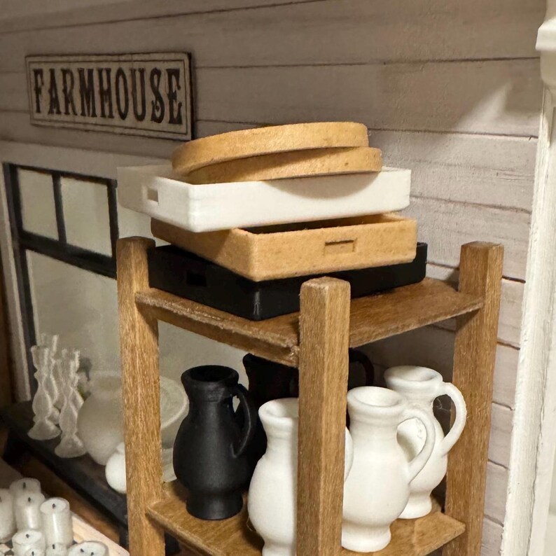 May include: A wooden shelf unit with three shelves, holding a stack of three trays, two white and one brown, and a collection of white and black ceramic pitchers. The top shelf has a sign that reads "FARMHOUSE".