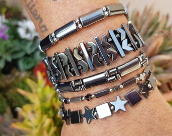 Puede incluir: Un conjunto de cinco pulseras de hematites negras con diferentes diseños, que incluyen un patrón geométrico, una fila de cuentas rectangulares y un diseño de estrella.