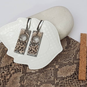 May include: A pair of dangle earrings with a snake skin pattern and silver eye charms. The earrings are displayed on a white plate with a brown and white patterned background.