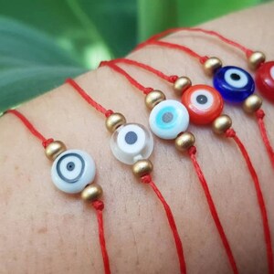 May include: Six red string bracelets with different coloured evil eye charms. The charms are blue, red, white, and turquoise. The bracelets are tied on a person's wrist.