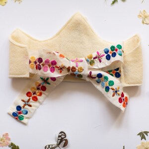 May include: A white felt crown with a colorful embroidered ribbon tied in a bow. The ribbon features a floral pattern in shades of pink, blue, green, yellow, and red.