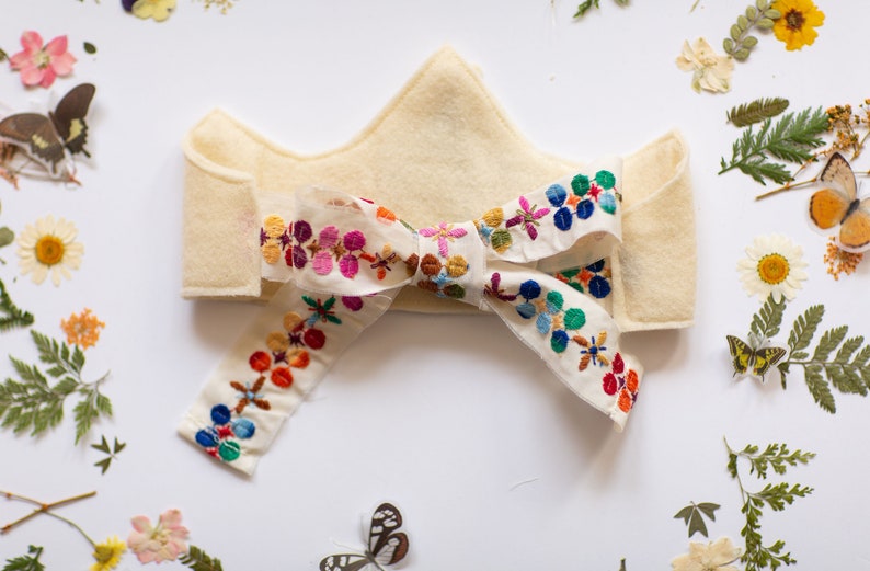 May include: A white felt crown with a colorful embroidered ribbon tied in a bow. The ribbon features a floral pattern in shades of pink, blue, green, yellow, and red.