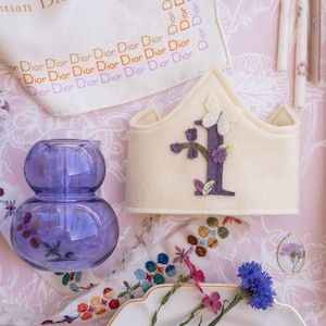 May include: A white felt crown with a purple felt letter I and purple felt flowers. The crown is on a pink and white floral patterned surface with a clear glass vase and a white plate with flowers.