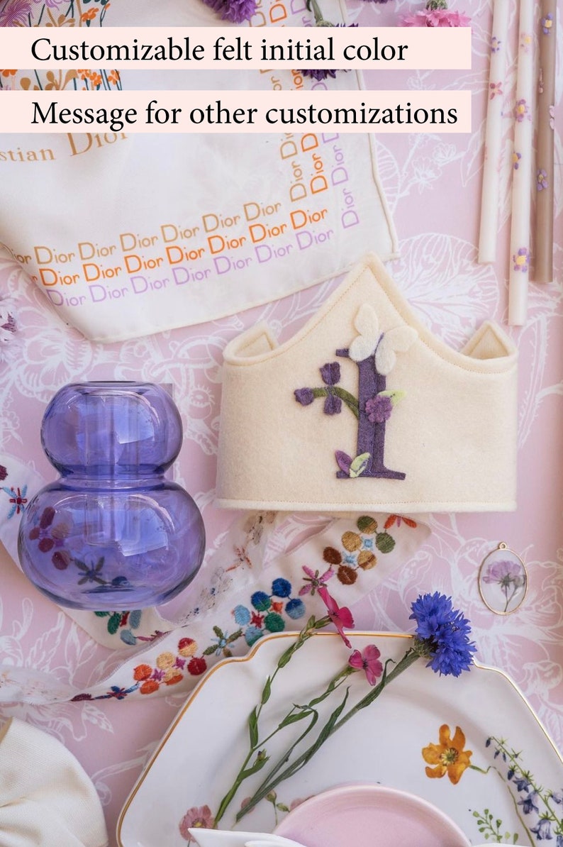 May include: A white felt crown with a purple felt letter I and purple felt flowers. The crown is on a pink and white floral patterned surface with a clear glass vase and a white plate with flowers.