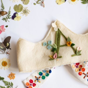 May include: A white felt crown with a green felt letter A decorated with colorful felt flowers. The crown is paired with a white fabric band with colorful embroidery.
