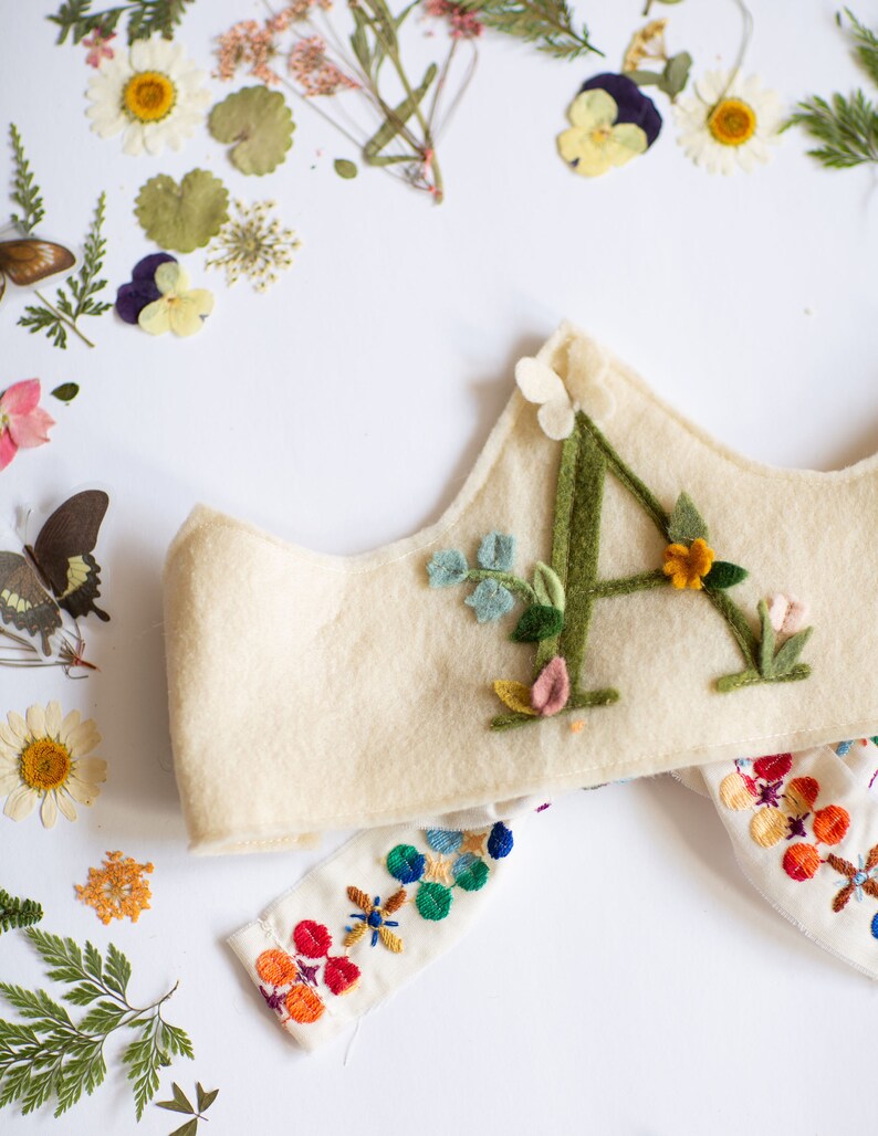 May include: A white felt crown with a green felt letter A decorated with colorful felt flowers. The crown is paired with a white fabric band with colorful embroidery.