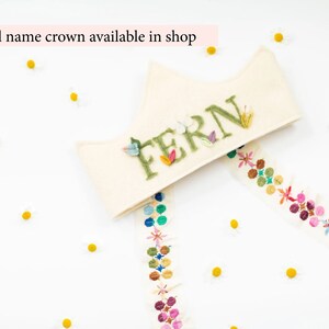 May include: A white felt crown with the name "FERN" embroidered in green letters with green leaves. The crown is decorated with a colorful ribbon with embroidered flowers.
