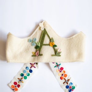 May include: A white felt crown with a green felt letter A embroidered on the front. The letter A is surrounded by green felt leaves and colorful felt flowers. The crown has two white felt ties with colorful embroidered flowers.