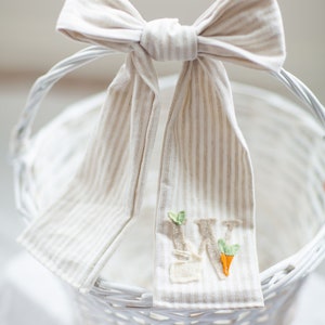 May include: White wicker basket with a beige and white striped fabric bow tied around the handle. The fabric has an embroidered letter "W" with green leaves and an orange carrot.