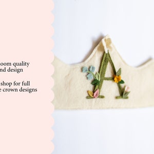 May include: A felt crown with the letter A embroidered in green felt and surrounded by small felt flowers in shades of pink, yellow, and green.