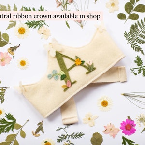 May include: A felt crown with a green felt letter A embroidered on it. The crown is decorated with small felt flowers and leaves. The crown is tied with a white ribbon.