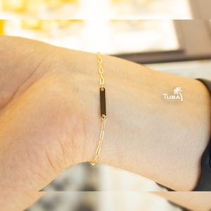 May include: A gold-colored chain bracelet with a rectangular black accent piece. The bracelet is worn on a wrist, and the chain links are small and delicate. The bracelet is a piece of jewelry.