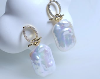 Pode incluir: Um par de brincos de ouro com uma gota de pérola e um pequeno círculo de pedras brilhantes. Os brincos estão sobre uma superfície branca.