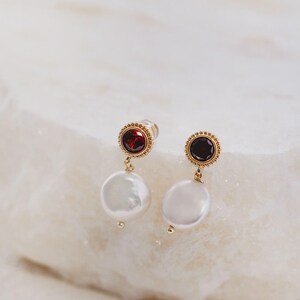 May include: A pair of gold-tone drop earrings featuring a circular stud with a deep red garnet-colored stone surrounded by a beaded border. A white, flat, coin-shaped pearl hangs from each stud, accented by a small gold-tone bead at the bottom.