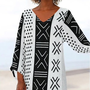 May include: A white tunic top with a V-neck and long sleeves. The top features a bold black and white geometric pattern, including dots, lines, and cross-like shapes. The design is symmetrical, with the pattern repeating on both sides of the top.