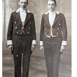 May include: A vintage black and white photograph of two men in formal attire. They are wearing dark jackets with white lapels and cuffs, white bow ties, and vests. The background is a simple, light-colored wall.