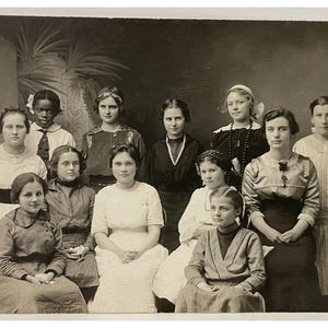 May include: A vintage black and white photograph featuring a group of women. They are dressed in various styles of clothing, including dresses, blouses, and skirts. The women are posed in front of a dark backdrop, with some seated and others standing.