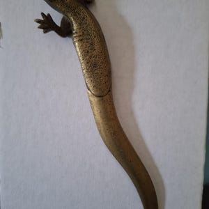 May include: A metallic lizard-shaped object with a textured, bronze-toned finish. The figure features a detailed head, four splayed legs with visible toes, and a long, smooth, tapering tail that curves slightly towards the bottom of the frame.