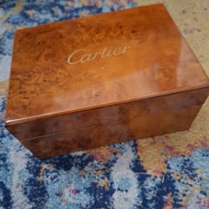 Puede incluir: Una caja rectangular de madera con un acabado pulido y brillante que presenta un patrón de veta de madera nudosa en tonos marrón y ámbar. El nombre de la marca Cartier está grabado en letras doradas en el centro de la tapa de este contenedor.