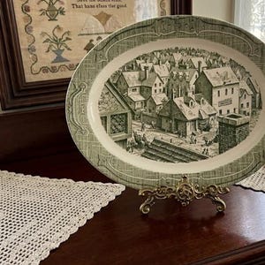May include: Oval serving platter with a green and white design. The center features a detailed illustration of a town scene. The platter is displayed on a decorative gold-colored stand, showcasing a vintage aesthetic.