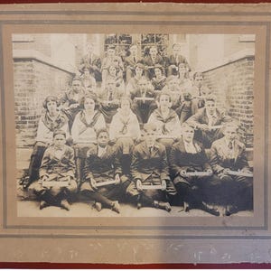 May include: A vintage black and white photograph of a group of people, likely a class or organization, posed on steps. The subjects are dressed in formal attire, with some holding what appear to be diplomas or certificates. The photograph is framed with a decorative border.