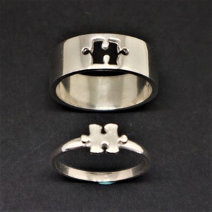 May include: Two silver puzzle piece rings. The top ring has a black puzzle piece cutout. The bottom ring has a silver puzzle piece.