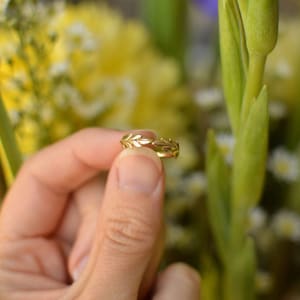 May include: A gold ring with a leaf design is held up against a blurred background of yellow and green flowers. The ring's intricate details are highlighted in this close-up shot. The ring is a piece of jewellery.