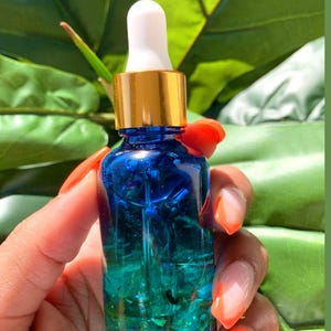May include: A small, cylindrical blue glass bottle featuring a gold-colored collar and a white rubber dropper top. The bottle is filled with a clear blue liquid containing visible green and blue glitter particles throughout the base.