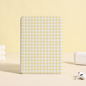 Puede incluir: Una funda con estampado de cuadros amarillos y blancos está en posición vertical. Una funda de carga blanca y un par de auriculares inalámbricos blancos están cerca. Una vela blanca con forma de burbuja está en el fondo.