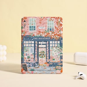 Puede incluir: Una funda rectangular con una ilustración de una librería, una bicicleta y hojas de otoño. La tienda tiene un toldo azul y las palabras "THE BOOKSHOP" sobre la entrada. La funda está sobre una superficie amarillo claro.