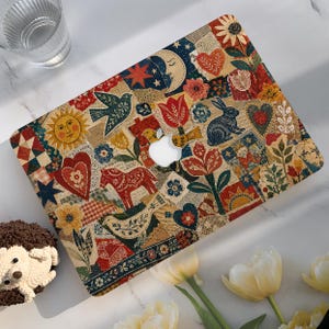 Könnte beinhalten: Eine Laptop-Hülle mit einer farbenfrohen Volkskunst-Collage aus Motiven von Sonne, Mond, Hase, Pferd, Vögeln, Herzen und Blumen in Rot-, Blau-, Gelb- und Grüntönen, inklusive einer zentralen Aussparung für das Apple-Logo.