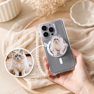Puede incluir: Una funda transparente para smartphone con un accesorio magnético circular que muestra la imagen impresa de un gato blanco esponjoso de ojos azules. La funda incluye una correa de muñeca de perlas blancas a juego con la foto del gato.