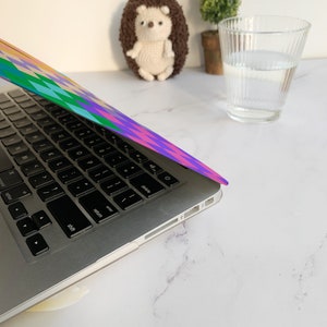 K&ouml;nnte beinhalten: Ein silberner Laptop mit einem bunten geometrischen Muster auf dem Deckel. Der Laptop ist ge&ouml;ffnet und die Tastatur ist sichtbar. Der Laptop steht auf einer wei&szlig;en Oberfl&auml;che.