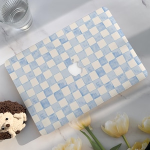 May include: A laptop featuring a light blue and white chequered design. The Apple logo is visible. The laptop is accompanied by a glass of water, a hedgehog pastry, and yellow tulips.