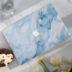May include: A laptop with a blue and white marble design. The laptop has a white Apple logo in the center. The laptop is on a white surface with a glass and flowers.