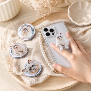 Può includere: Una collezione di supporti circolari per telefono e un supporto fissato a una custodia trasparente per smartphone, ognuno con l'immagine stampata di un gatto bianco soffice con occhi blu. Un cinturino di perle bianche è fissato alla custodia.