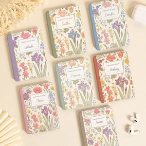 May include: Seven rectangular composition books with floral covers featuring various colored spines. Each book has a white label with the text Composition Book and a unique name: Belinda, Caitlin, Deirdre, Elaine, Frances, Hedwig, and Karen.