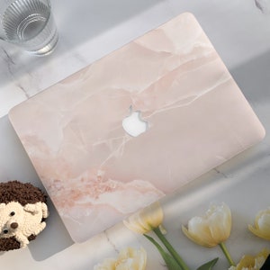 Puede incluir: Una piel de portátil de mármol rosa con un logotipo de Apple plateado. La piel tiene un fondo rosa claro con vetas blancas.