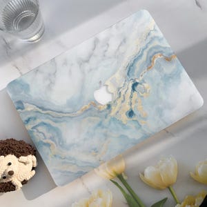 May include: A laptop case featuring a marble effect in blue, white, and gold. The case has a cut-out for the Apple logo. The rectangular case has rounded corners. The case is on a white surface.