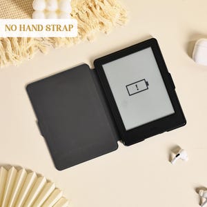 Może przedstawiać: Czarny czytnik e-book&oacute;w w dopasowanym czarnym etui, pokazany w pozycji otwartej. Na ekranie widoczna jest ikona niskiego poziomu baterii z wykrzyknikiem. W lewym g&oacute;rnym rogu znajduje się napis NO HAND STRAP. Urządzenie ma smukłą, prostokątną ramkę.