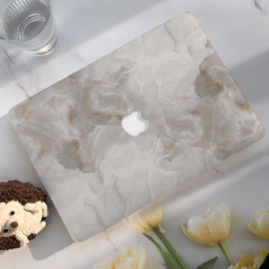 Può includere: Una pelle per laptop bianca con un motivo a marmo e un logo Apple argentato. La pelle è su una superficie bianca con un piccolo riccio di peluche e fiori gialli.