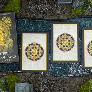 May include: A deck of "The Esoteric Buddhism of Japan Oracle Cards" by Yuzui Kotaki, with three cards showing a detailed wheel design. The book cover features a golden Buddha image. The cards are set on a textured surface.